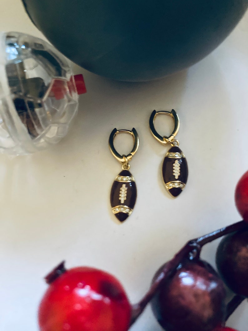 May include: A pair of gold hoop earrings with a brown football charm. The football charm has white stitching and a small diamond accent.