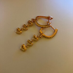May include: A pair of gold hoop earrings with a dangling chain of five small heart charms. Each heart charm is encrusted with tiny clear stones.