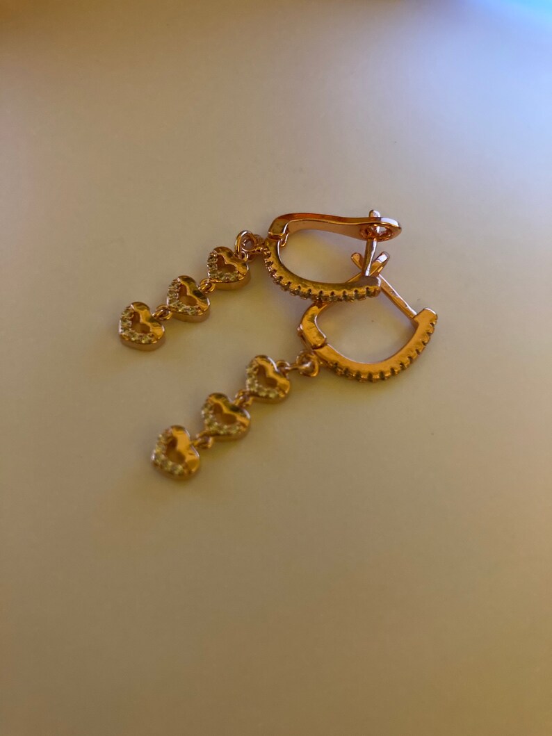 May include: A pair of gold hoop earrings with a dangling chain of five small heart charms. Each heart charm is encrusted with tiny clear stones.