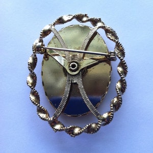 K&ouml;nnte beinhalten: Goldfarbene Brosche mit einem ovalen Design im Kameenstil. Das Oval ist von einem gedrehten seilartigen Rand umgeben. Die R&uuml;ckseite der Brosche hat eine Nadel und einen Verschluss.