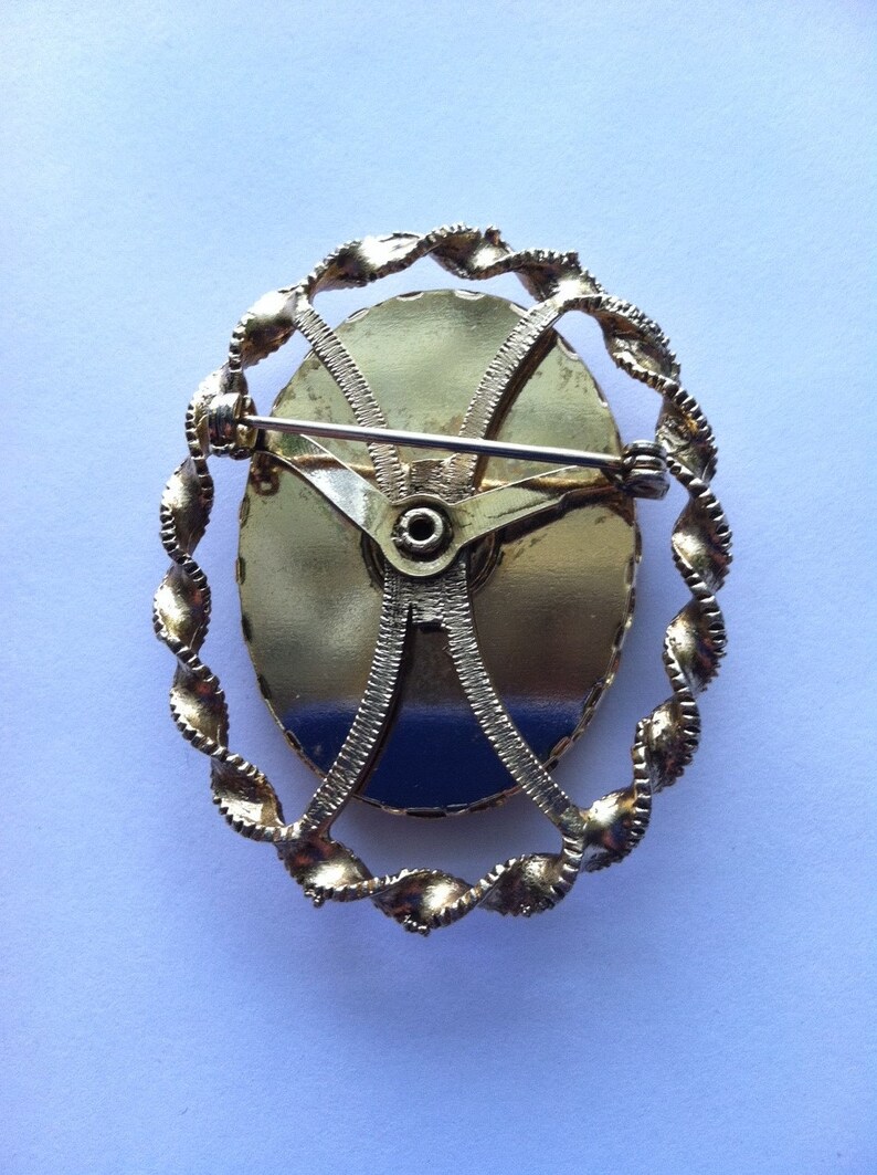 K&ouml;nnte beinhalten: Goldfarbene Brosche mit einem ovalen Design im Kameenstil. Das Oval ist von einem gedrehten seilartigen Rand umgeben. Die R&uuml;ckseite der Brosche hat eine Nadel und einen Verschluss.