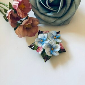 May include: A vintage porcelain brooch featuring a cluster of blue and white forget-me-not flowers with green leaves and pink accents.