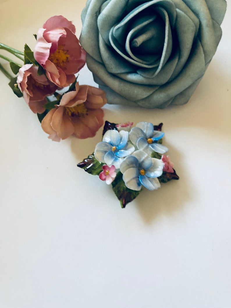 May include: A vintage porcelain brooch featuring a cluster of blue and white forget-me-not flowers with green leaves and pink accents.