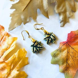 May include: Gold-toned leaf earrings with green and gold accents. The earrings are shaped like leaves and have a hook closure. The image also features decorative fall leaves in shades of gold, orange, and red.