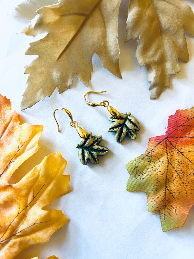 May include: Gold-toned leaf earrings with green and gold accents. The earrings are shaped like leaves and have a hook closure. The image also features decorative fall leaves in shades of gold, orange, and red.