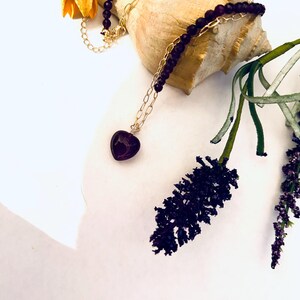 May include: A purple heart pendant necklace with a gold chain and purple beads. The necklace is displayed on a seashell and a sprig of purple flowers.