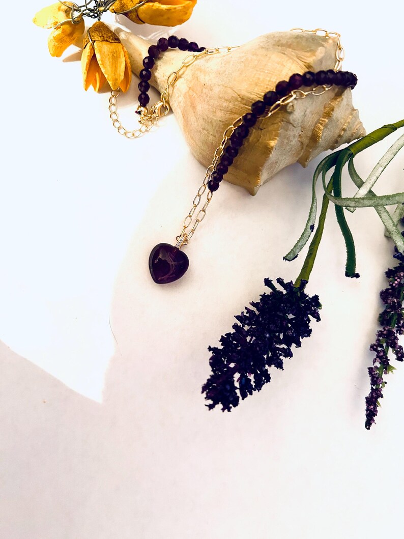 May include: A purple heart pendant necklace with a gold chain and purple beads. The necklace is displayed on a seashell and a sprig of purple flowers.