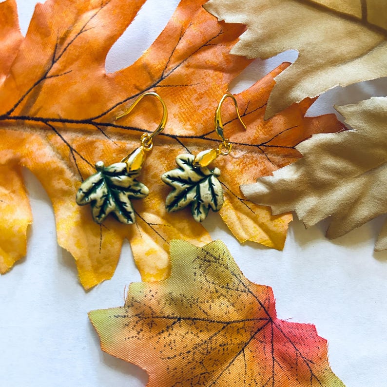 May include: A pair of gold-toned earrings featuring leaf-shaped pendants with green and cream detailing. The earrings are displayed on a backdrop of colorful autumn leaves in shades of orange, yellow, and brown, evoking a fall theme.