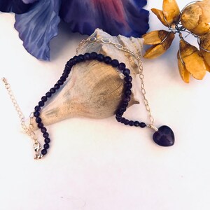 May include: A delicate gold chain necklace with a purple heart pendant and a strand of dark purple beads. The necklace is displayed on a seashell.