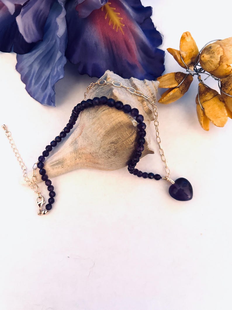 May include: A delicate gold chain necklace with a purple heart pendant and a strand of dark purple beads. The necklace is displayed on a seashell.