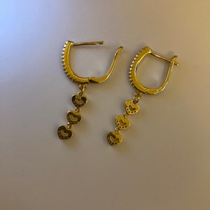 May include: A pair of gold hoop earrings with a dangling chain of four heart-shaped charms. Each heart charm is decorated with small, sparkling stones.