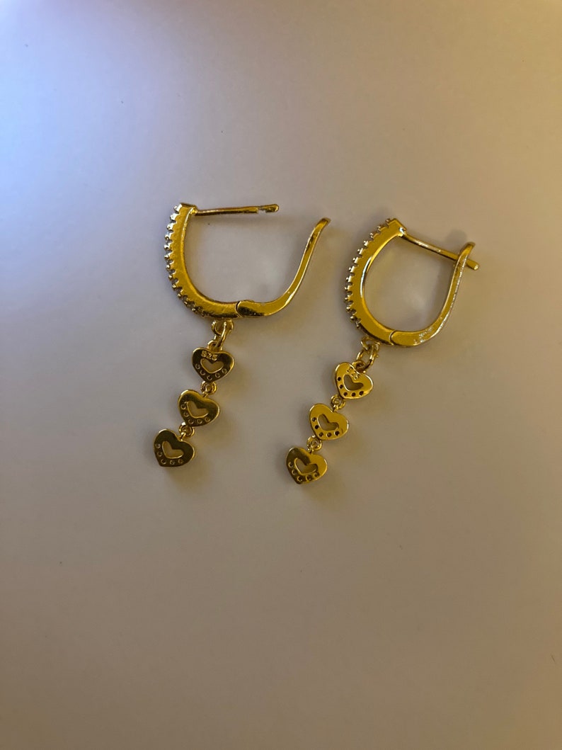 May include: A pair of gold hoop earrings with a dangling chain of four heart-shaped charms. Each heart charm is decorated with small, sparkling stones.