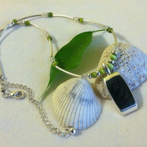 May include: A silver necklace with a green and white beaded chain and a dark green pendant. The pendant is a rectangular shape with a swirl design. The necklace is displayed on a white surface with two seashells.