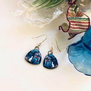 May include: A pair of gold-tone dangle earrings. Each earring features a blue, triangular pendant with a whimsical illustration of a white cat, a house, and trees. The earrings are set against a white background.