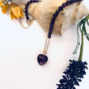 May include: A delicate gold chain necklace with a purple heart-shaped pendant. The necklace is made of small purple beads and is displayed on a seashell.