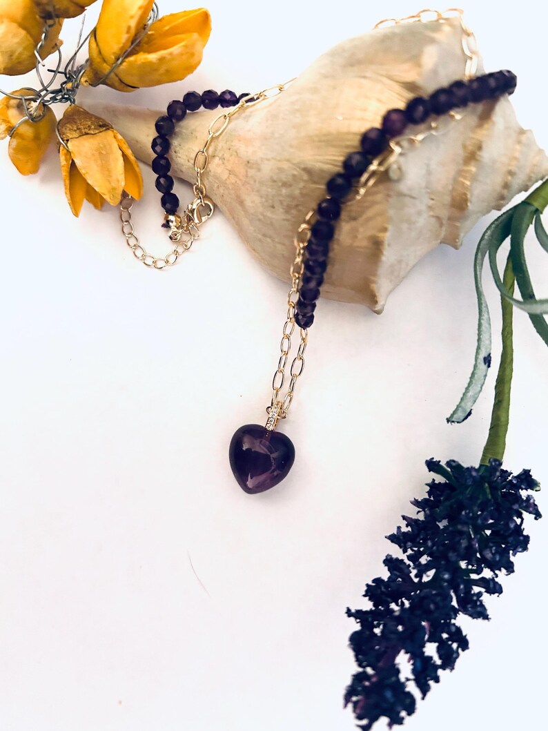 May include: A delicate gold chain necklace with a purple heart-shaped pendant. The necklace is made of small purple beads and is displayed on a seashell.