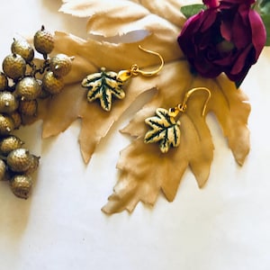 May include: Gold-toned leaf earrings with green and cream enamel detailing. The earrings are shaped like maple leaves and hang from gold-colored hooks. The earrings are displayed on a large, tan leaf with gold-colored berries and a burgundy flower.