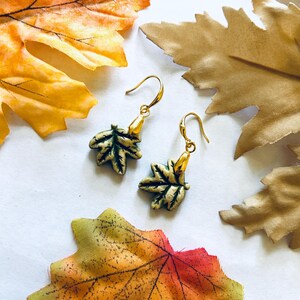 May include: Gold-colored leaf earrings with green and gold accents. The earrings are shaped like maple leaves and hang from gold-colored hooks. The background includes colorful fall leaves.