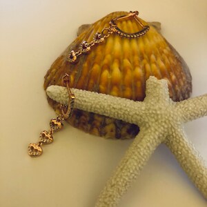 May include: A pair of gold-tone earrings with a heart-shaped design. The earrings are made of metal and have a dangling heart charm. The earrings are on a white background with a seashell and a starfish.