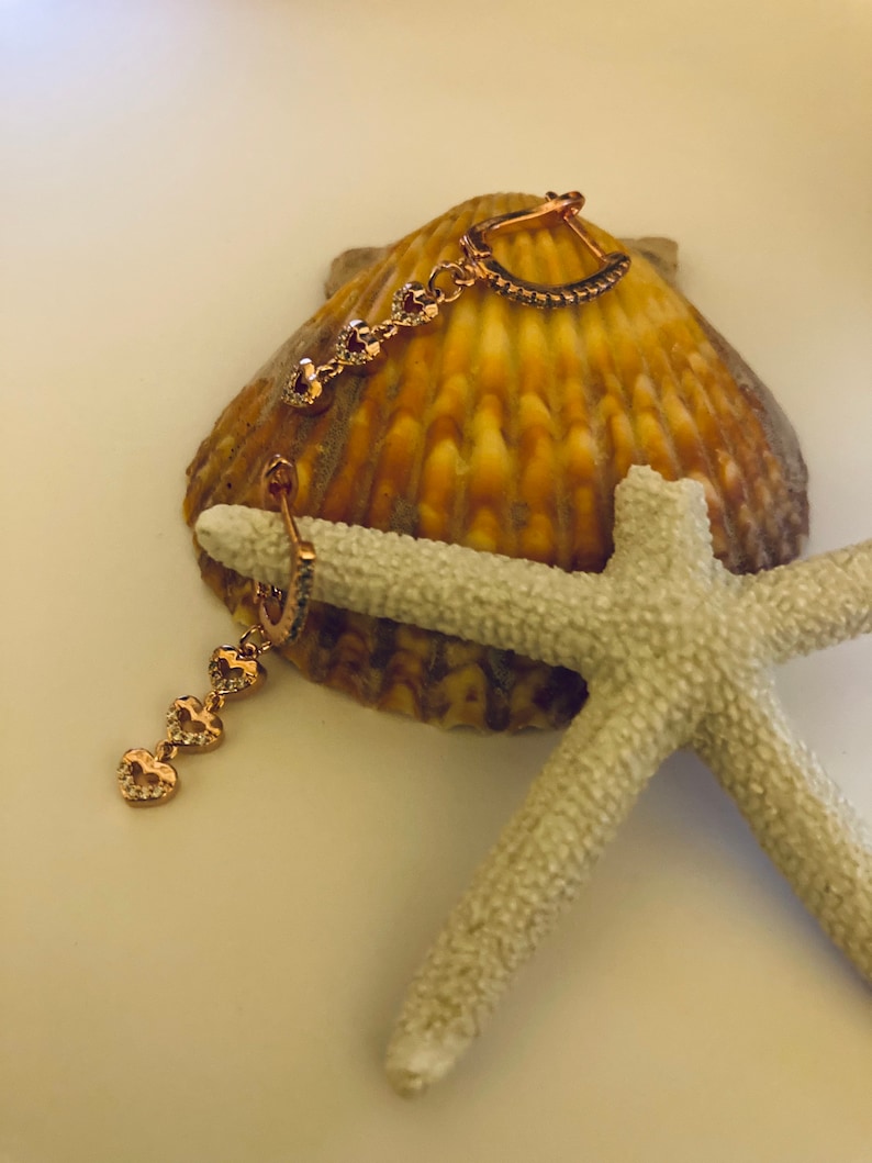 May include: A pair of gold-tone earrings with a heart-shaped design. The earrings are made of metal and have a dangling heart charm. The earrings are on a white background with a seashell and a starfish.