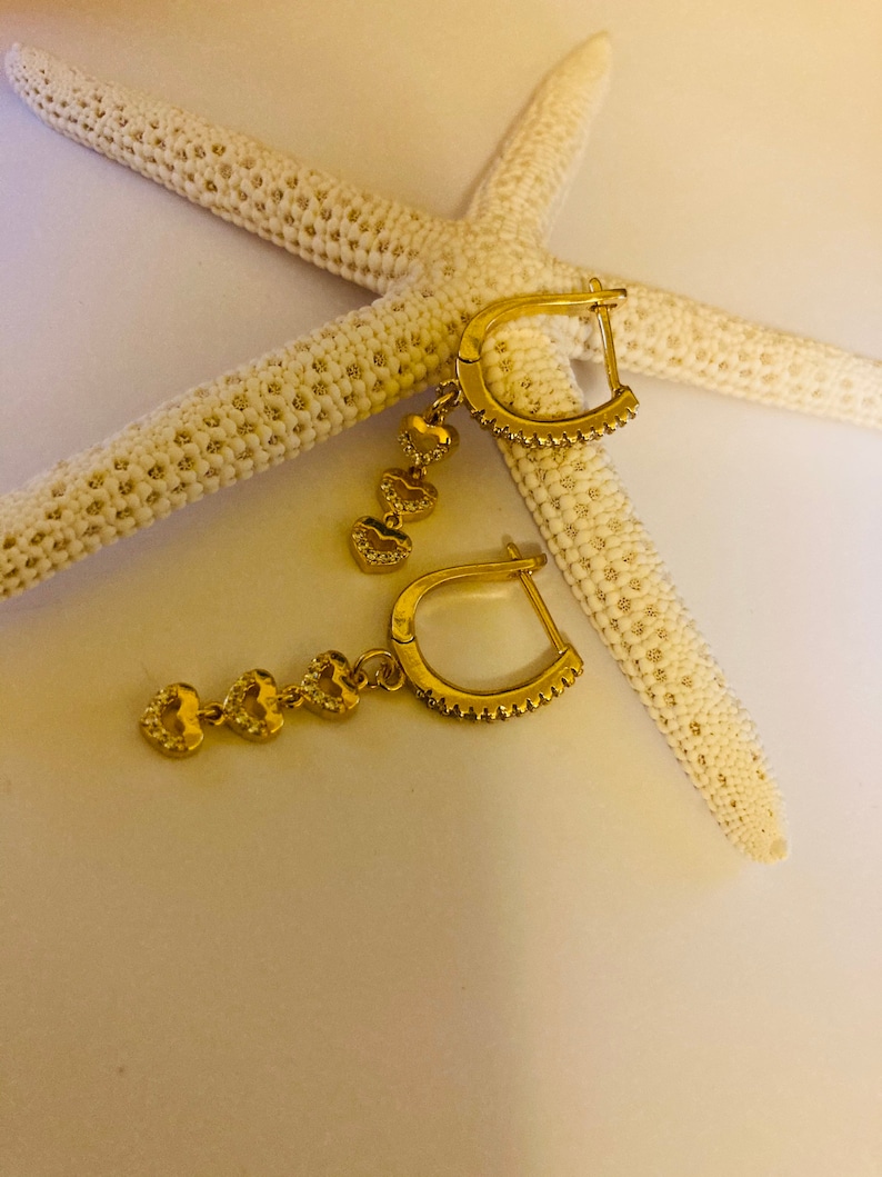 May include: A pair of gold hoop earrings with dangling heart charms. Each heart charm is adorned with small, clear cubic zirconia stones.