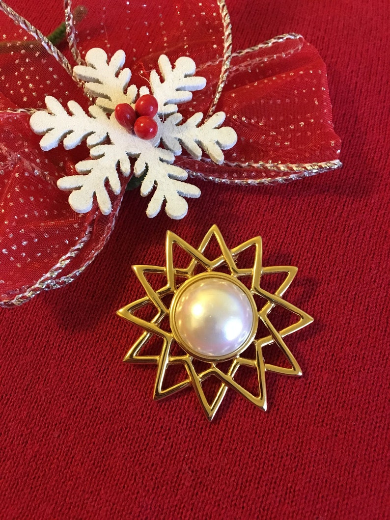 May include: A gold-toned starburst brooch with a large, pearlescent center stone. The brooch is set against a red background, near a white snowflake ornament with red berries and a red ribbon with silver trim. Festive and decorative.