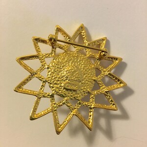 May include: Gold-toned starburst brooch with a textured center and a pin closure. The brooch features a detailed, openwork design with pointed rays. The back of the brooch has the initials "OAK" stamped on it.
