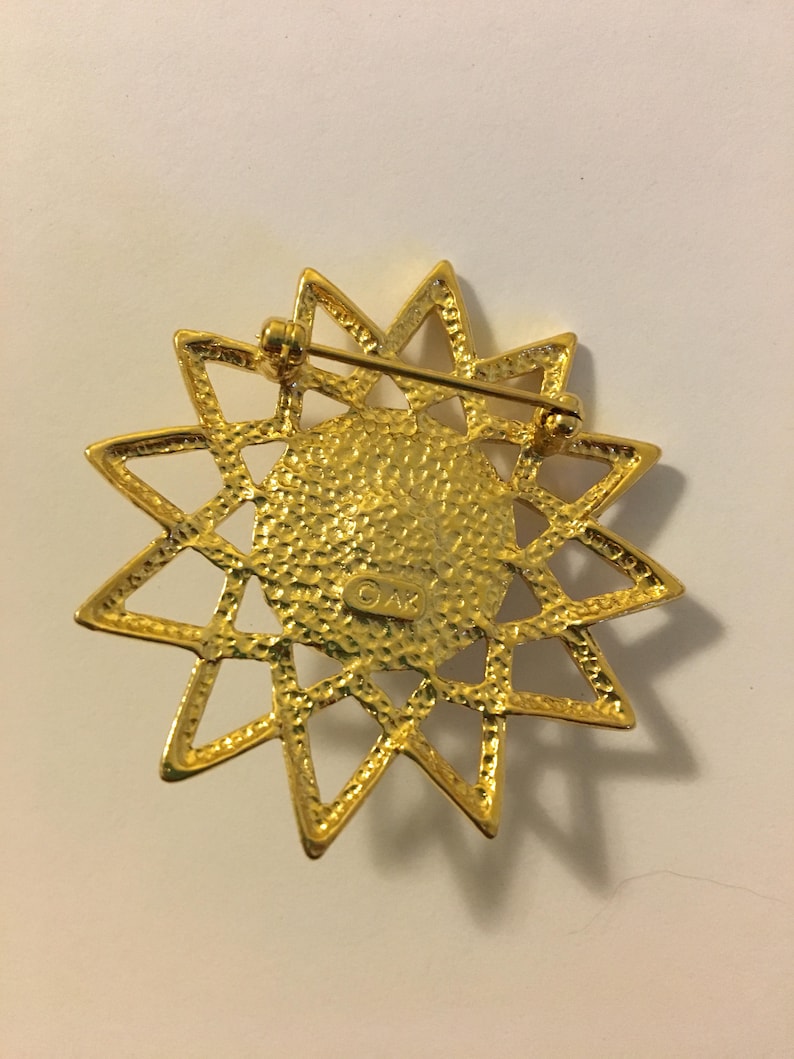 May include: Gold-toned starburst brooch with a textured center and a pin closure. The brooch features a detailed, openwork design with pointed rays. The back of the brooch has the initials "OAK" stamped on it.
