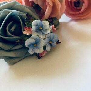 May include: A blue fabric rose with a cluster of blue and pink enamel flowers. The flowers are arranged in a bouquet and have a vintage style.