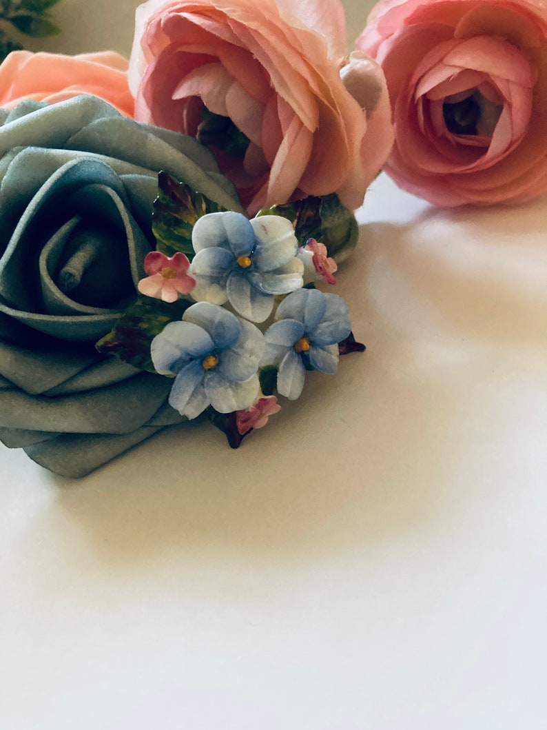 May include: A blue fabric rose with a cluster of blue and pink enamel flowers. The flowers are arranged in a bouquet and have a vintage style.