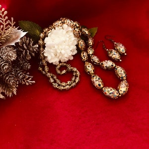 Puede incluir: Un conjunto de collar y pendientes de oro con cuentas ovaladas decoradas con motivos florales de esmalte de colores. El collar y los pendientes est&aacute;n sobre un fondo rojo.