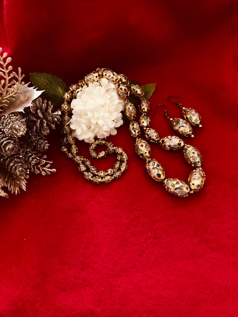 Puede incluir: Un conjunto de collar y pendientes de oro con cuentas ovaladas decoradas con motivos florales de esmalte de colores. El collar y los pendientes est&aacute;n sobre un fondo rojo.