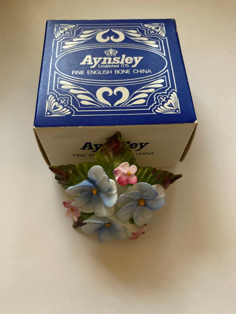 May include: A blue and white porcelain flower bouquet with green leaves. The bouquet is in a box that says "Aysnley Established 1775 Fine English Bone China".