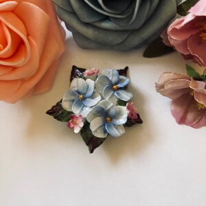 May include: A porcelain flower brooch with blue, white, and pink flowers with green leaves.