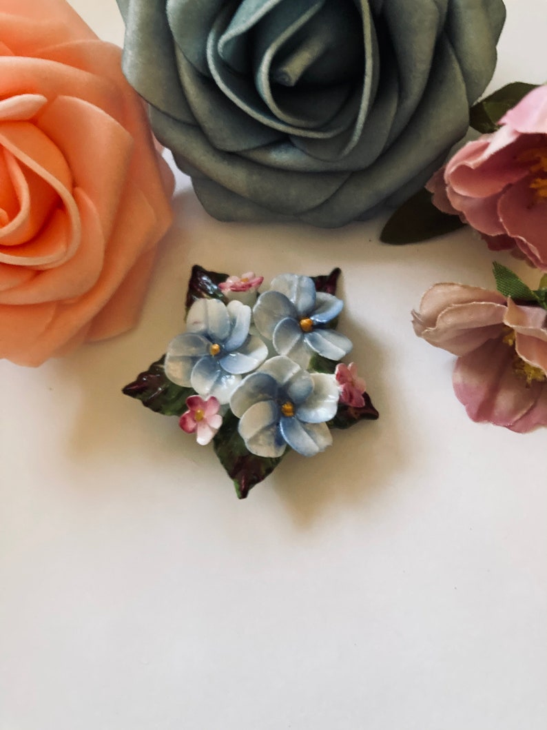 May include: A porcelain flower brooch with blue, white, and pink flowers with green leaves.