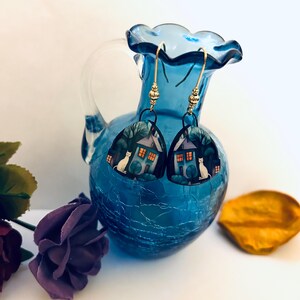 May include: A pair of dangle earrings featuring a whimsical house design with a white cat, trees, and a glowing window. The earrings are suspended from gold-colored hooks and are displayed against a blue vase.