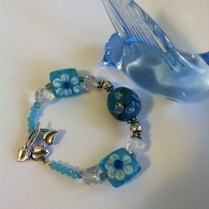 May include: A blue and silver bracelet with flower and bird charms. The bracelet is made with glass beads and silver accents.