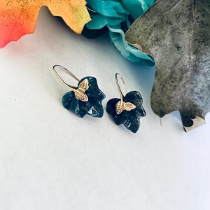 May include: A pair of dark green leaf-shaped earrings with gold butterfly accents. The earrings are dangling and have a silver hook.
