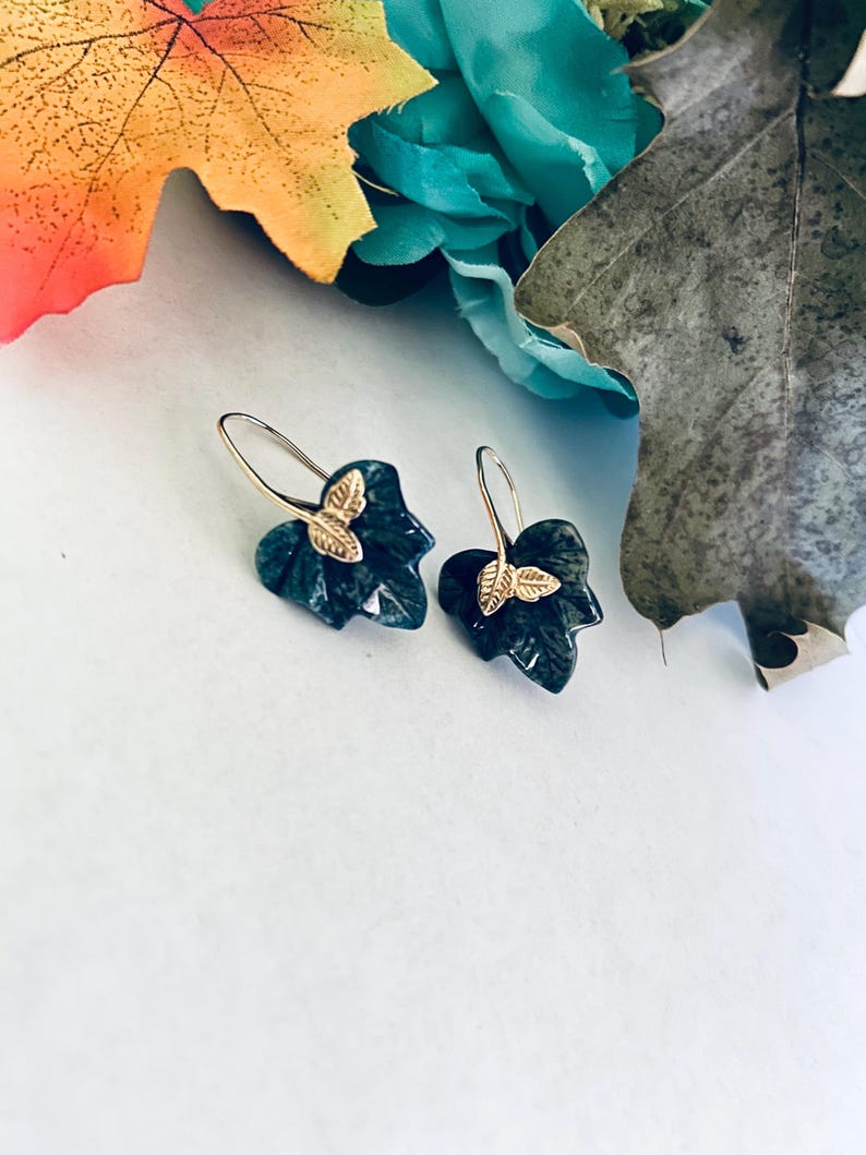 May include: A pair of dark green leaf-shaped earrings with gold butterfly accents. The earrings are dangling and have a silver hook.