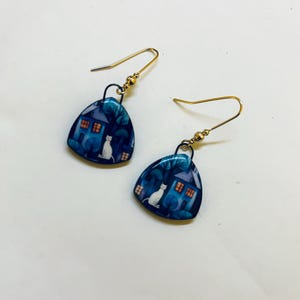 May include: A pair of gold-colored dangle earrings. Each earring features a blue, triangular charm with a white cat, a house, and trees. The earrings have a hook closure.