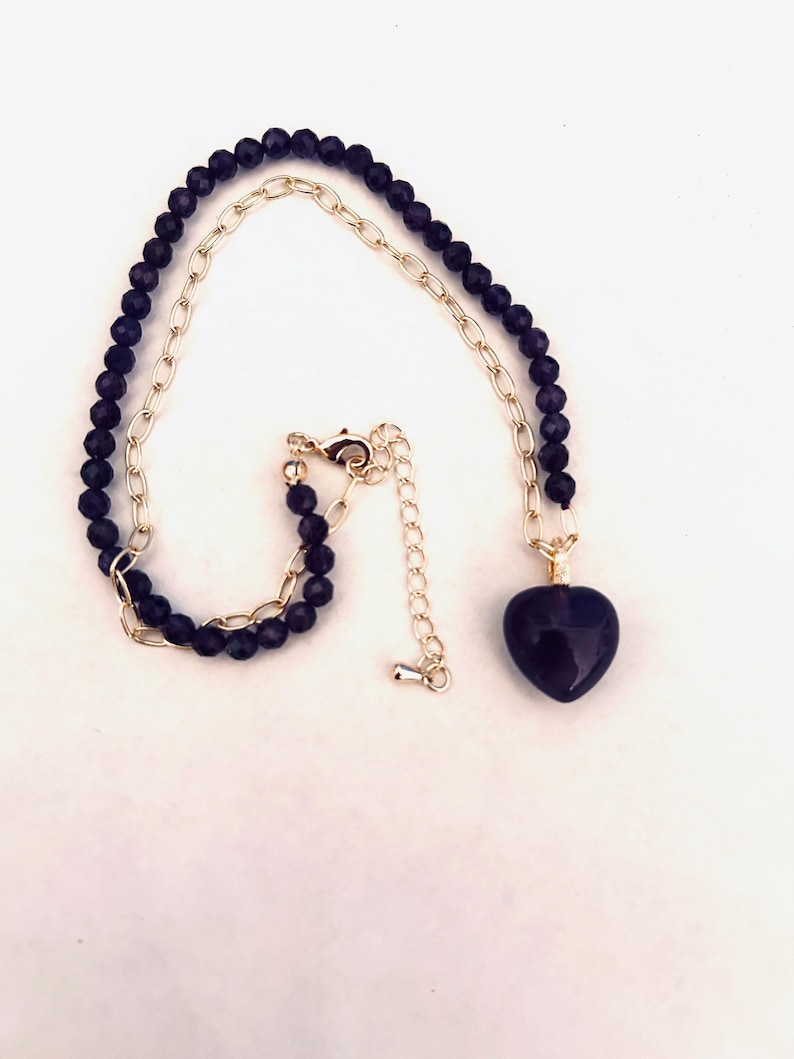 May include: A gold chain necklace with a dark purple heart pendant. The chain is made of small, round beads.