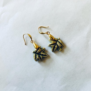May include: A pair of gold-colored leaf earrings with a hook closure. The earrings have a detailed leaf design with green and gold accents. The earrings are displayed on a white background.