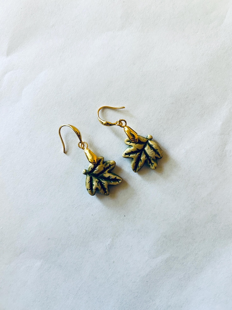 May include: A pair of gold-colored leaf earrings with a hook closure. The earrings have a detailed leaf design with green and gold accents. The earrings are displayed on a white background.