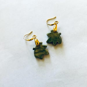 May include: A pair of leaf-shaped earrings. The earrings feature a dark green leaf design with gold-colored hooks. The earrings are set against a white background. The earrings are a jewelry item.