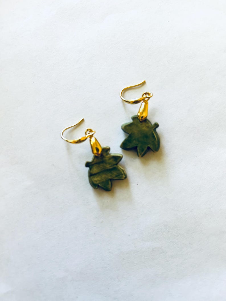 May include: A pair of leaf-shaped earrings. The earrings feature a dark green leaf design with gold-colored hooks. The earrings are set against a white background. The earrings are a jewelry item.
