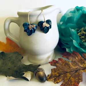 May include: A pair of dangle earrings with a teal blue leaf design and gold accents. The leaves are textured and have a realistic look.