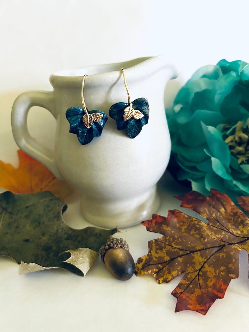 May include: A pair of dangle earrings with a teal blue leaf design and gold accents. The leaves are textured and have a realistic look.