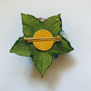 May include: A porcelain flower brooch with a green leaf design and a gold pin. The back of the brooch has the text "Made in England" and "Fine Bone China" in a gold circle.