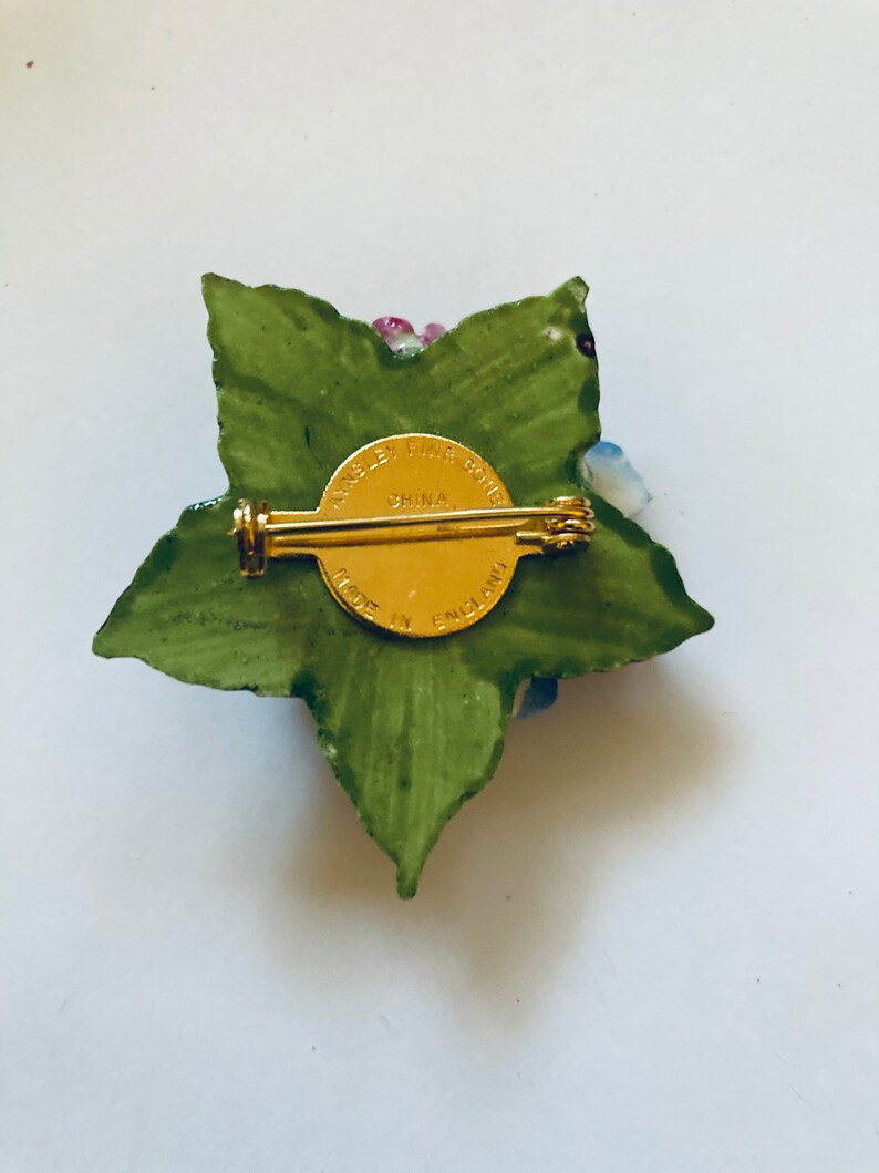 May include: A porcelain flower brooch with a green leaf design and a gold pin. The back of the brooch has the text "Made in England" and "Fine Bone China" in a gold circle.