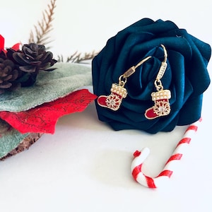 May include: Gold Christmas stocking earrings with red enamel and white pearl accents. The earrings are hanging from a blue velvet rose.